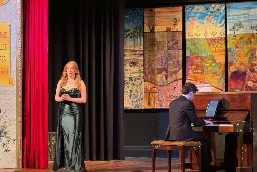 An opera singer stands on stage wearing a gown. She is near a pianist.