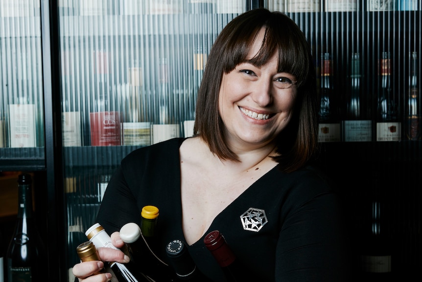 Woman with white skin and brown hair smiles holding wine bottles in front of a glass wine cabinet with bottles visible