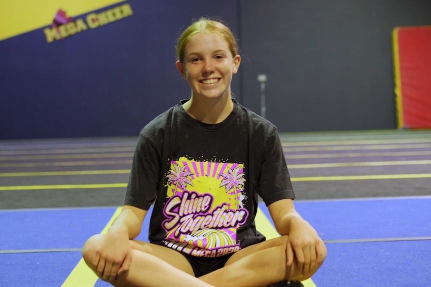 A girl in a cheerleading costume sits cross-legged looking at the camera