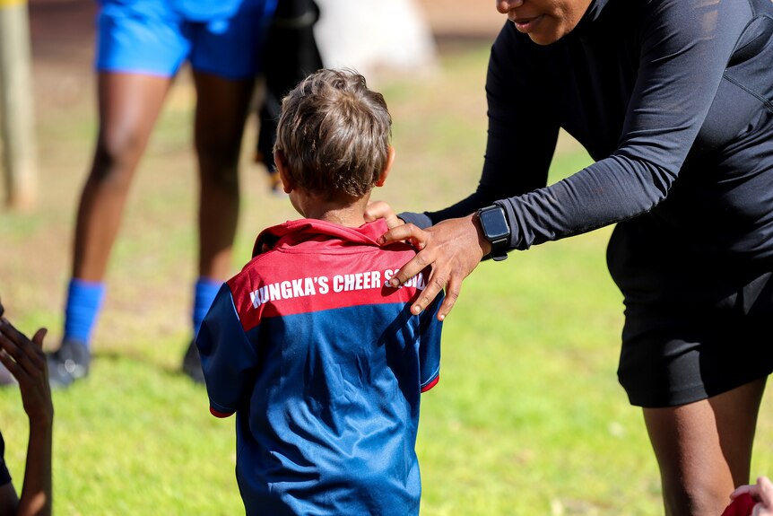 A young Aboriginal boy is helped by a woman with his shirt, which reads Kungka's Cheer Squad, while standing on football ground