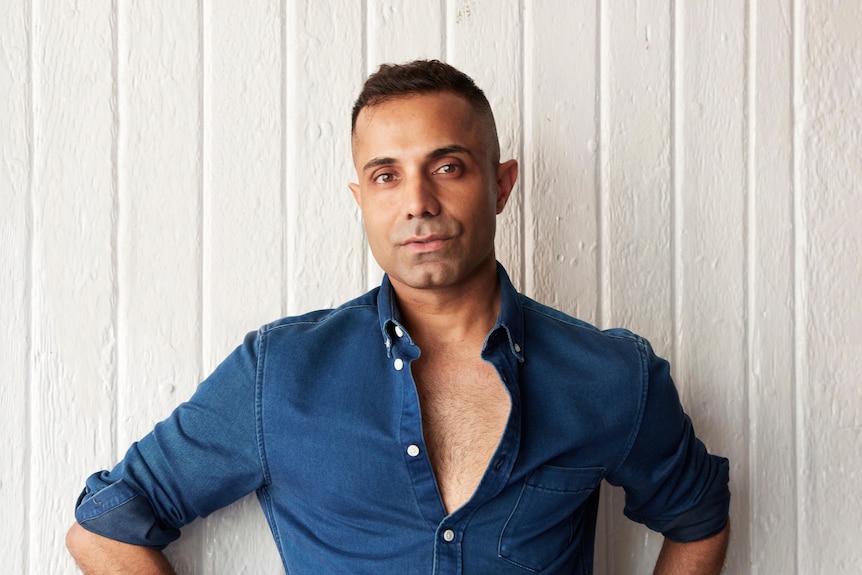 Indian-Australian man with short dark hair and brown eyes wears a denim blue shirt and stands in front of textured white wall.