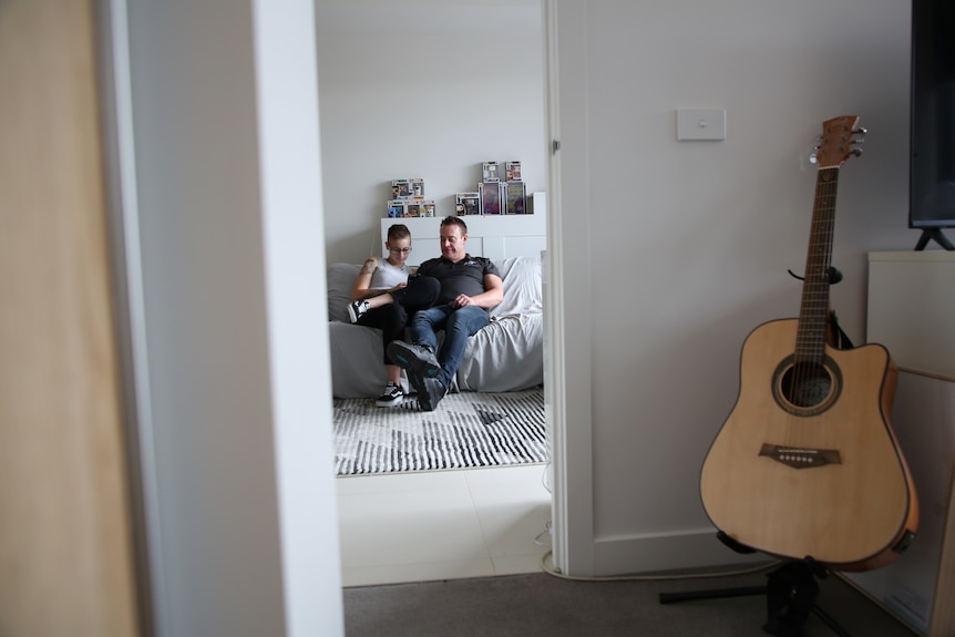 A man and a woman sit on a couch in their apartment in Melbourne, looking at the woman's phone.
