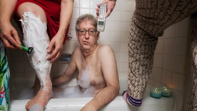 A man sitting in a bathtub looks nonplussed. A woman standing in the tub is shaving her legs and his mother's passing a phone