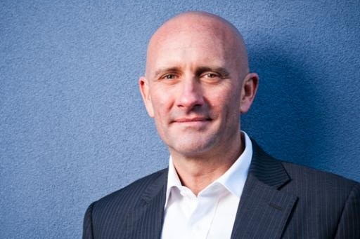 A man stands against blue wall wearing dark suit jacket and white shirt, smiling slightly with closed mouth.