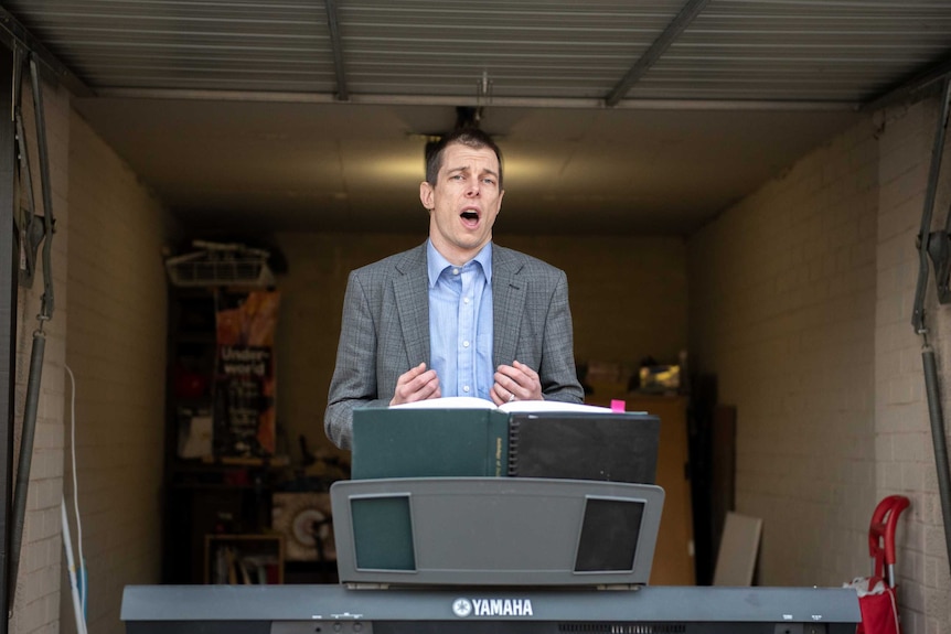 A man in a suit jacket sings opera in front of a keyboard, in the garage of his Sydney apartment.