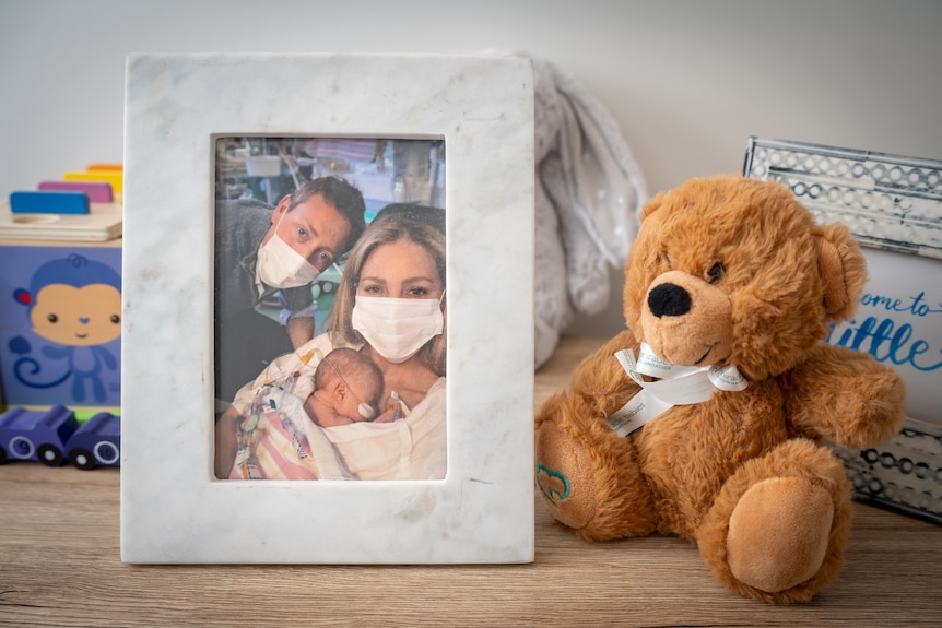 A photo of a women wearing a medical face mask and a premature baby with a stuffed teddy nearby