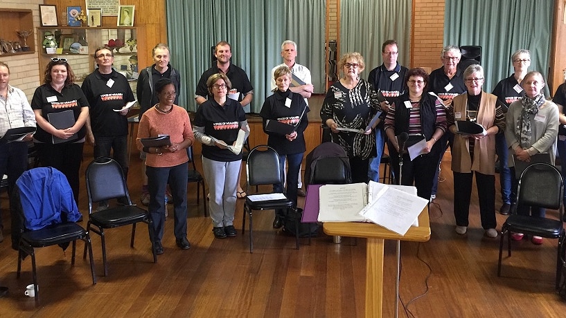 About 20 people standing, some holding books open, in a line as a choir.