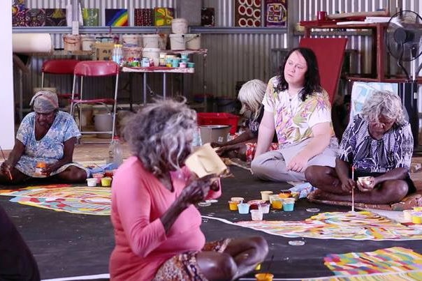 Anohni sits on the floor behind Martu elders as they paint a mural with paintbrushes.