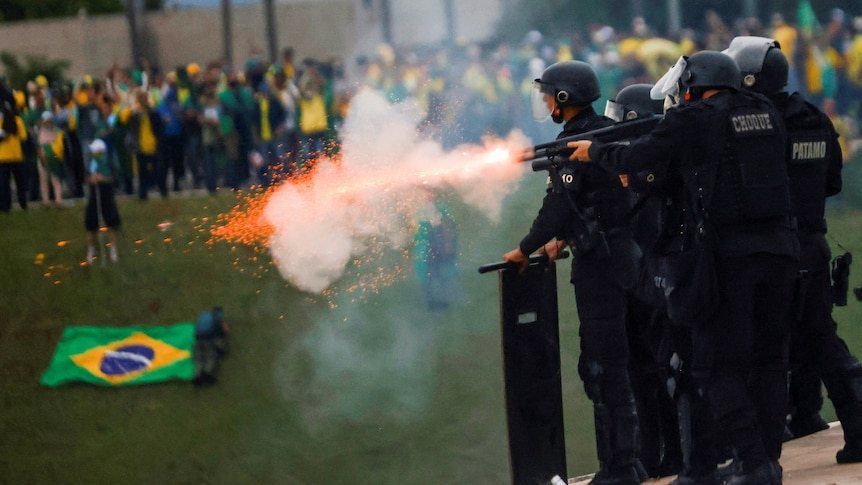 Sparks fly from a police officers gun as protesters in Brazilian flags crowd on a lawn. 