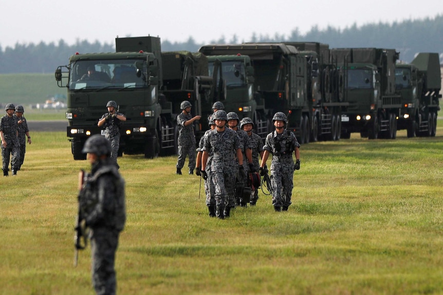Soldiers in a green field walk towards the camera with heavy military machinery in the background.