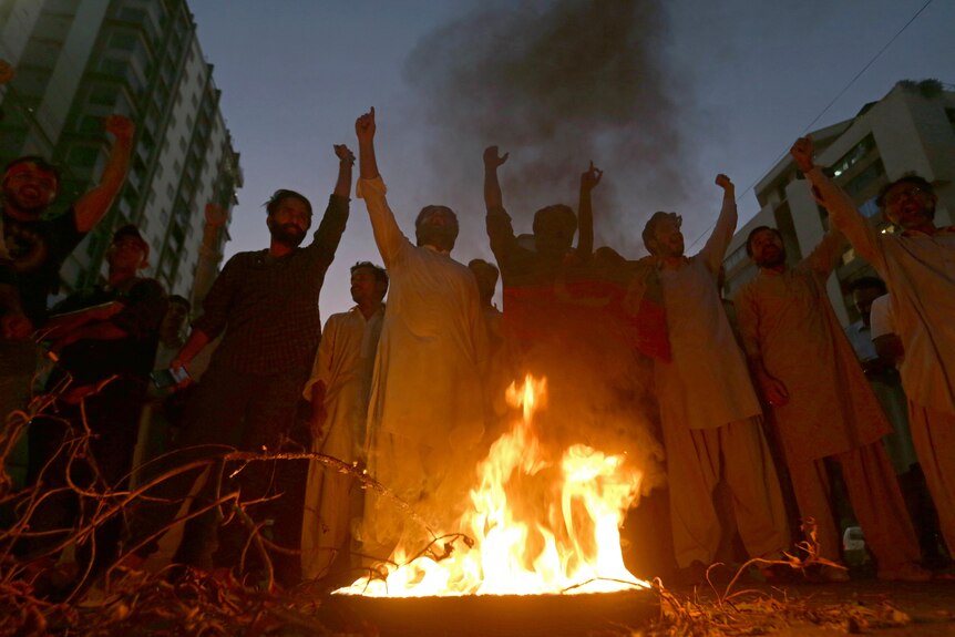 A group of political supporters stand around a small fire and raise their arms, with tall buildings behind.