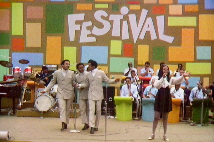 Black soul group Gladys Knight and the Pips perform enthusiastically in front of a brass band and colourful festival sign.
