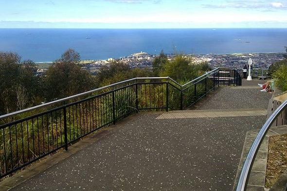 Vista of blue ocean and coastline in distance with fenced lookout and paved footpath in foreground