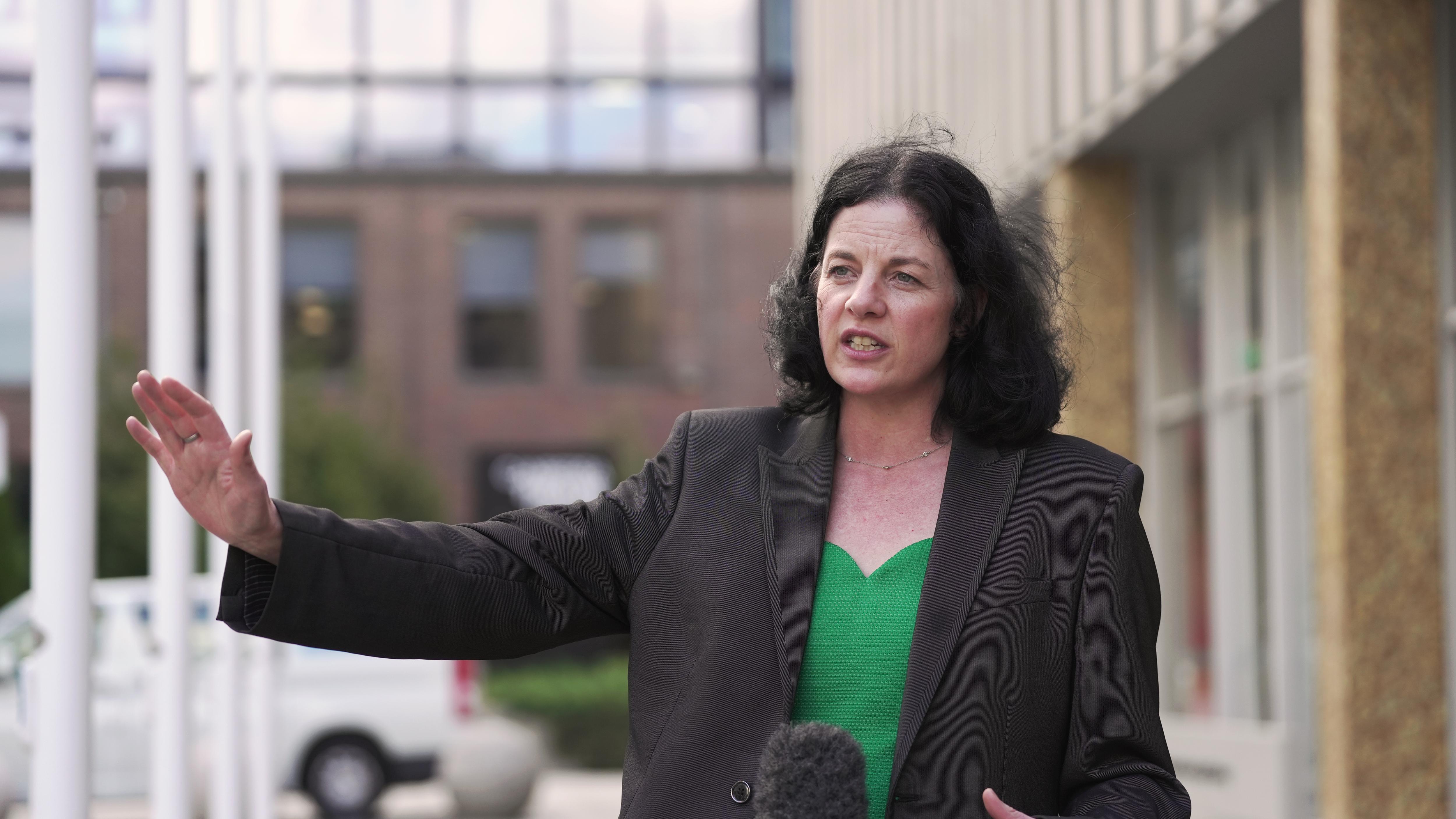 A woman with dark hair wearing a blazer gestures while speaking outside into microphone.