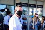 Mark McGowan wearing a black face mask and shirt with no tie, with a photographer in the background.
