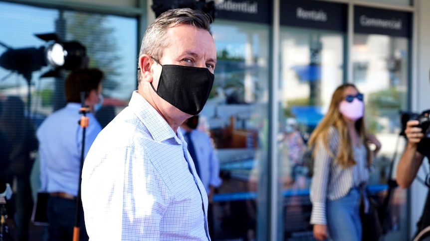 Mark McGowan wearing a black face mask and shirt with no tie, with a photographer in the background.