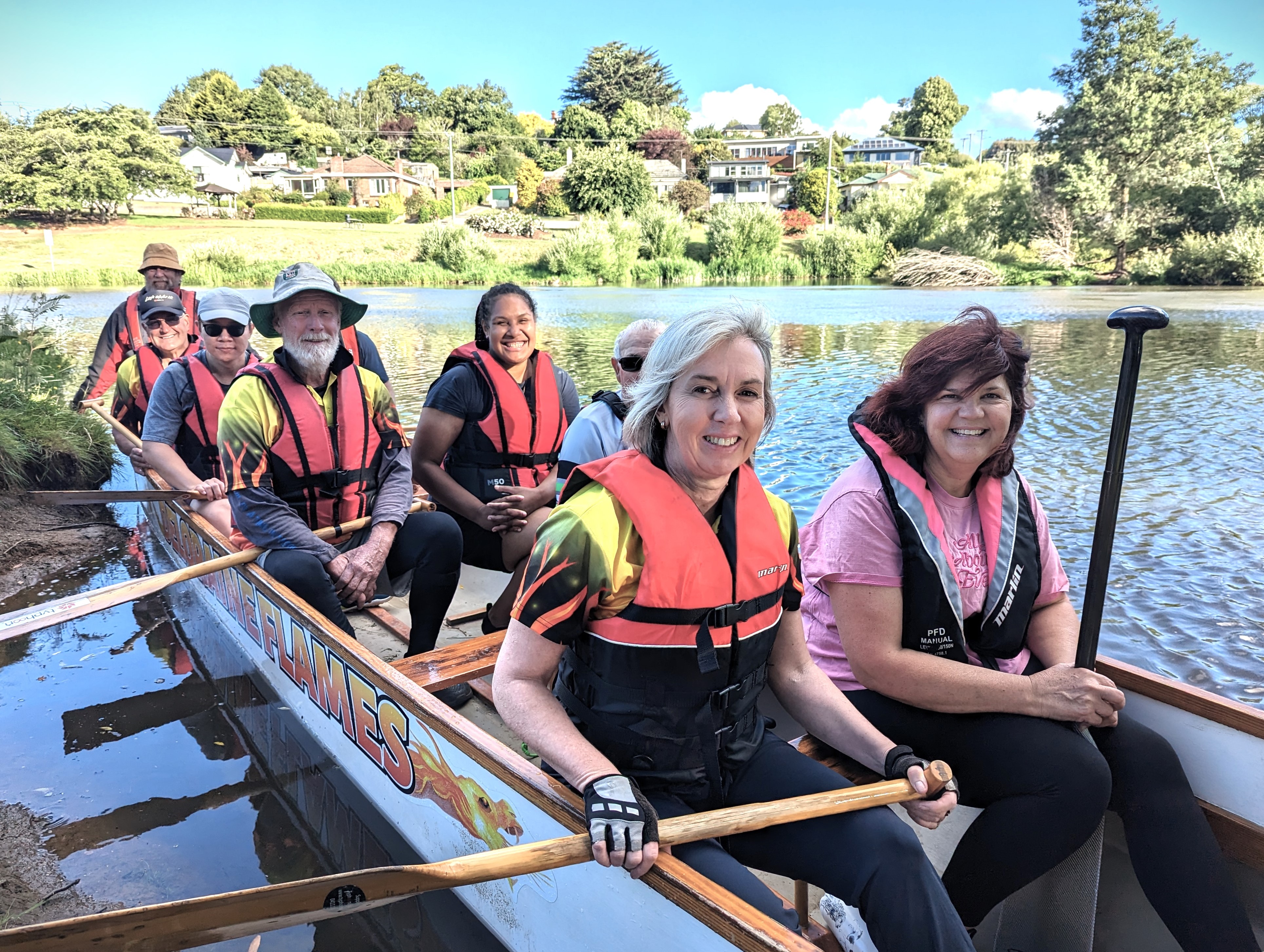 Eight people of different ages and walks of life sit in a dragon boat on a river, all smiling.