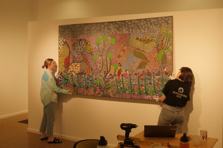 Two gallery workers lift a large painting into its place on the wall.