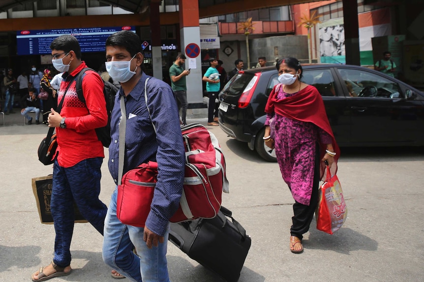 Indians arrive at a train station wearing masks and wheeling suitcases behind them.