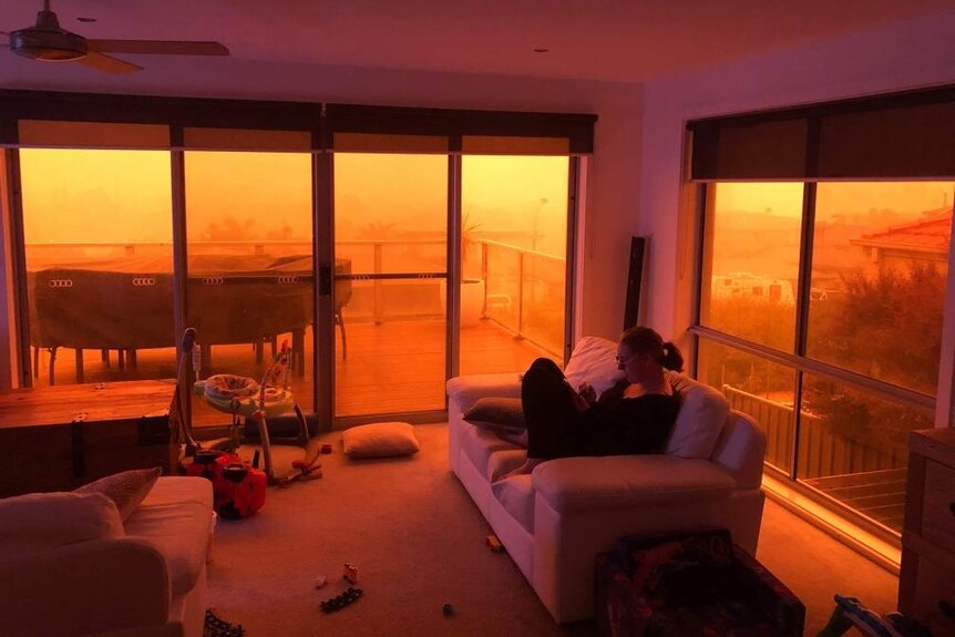A woman sits on her couch next to children's toys, with a view out her window of orange caused by bushfires.