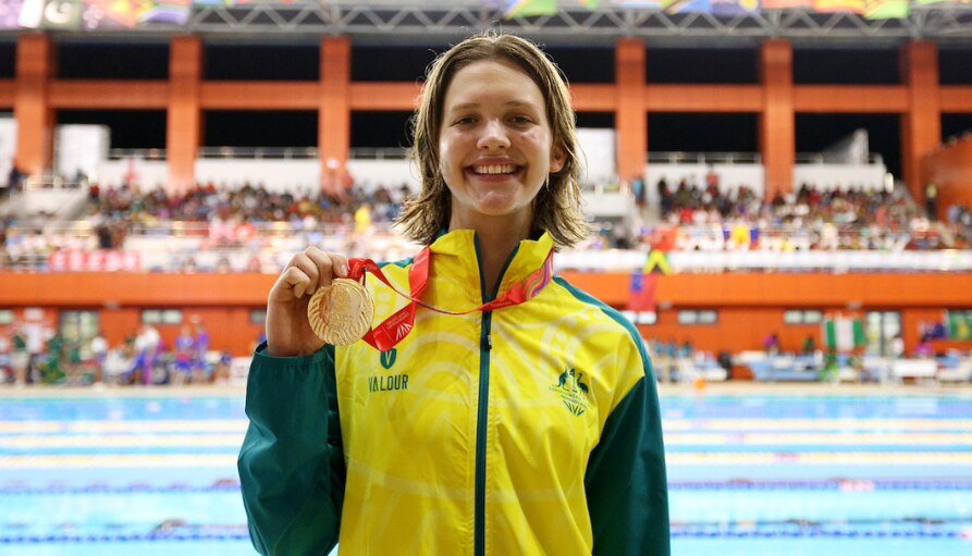 A young woman smiles in an Australian tracksuit while holding a medal that is around her neck.