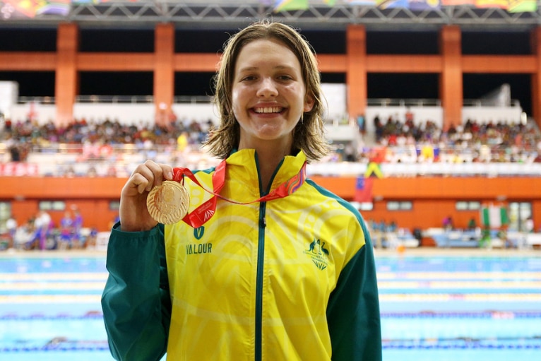 A young woman smiles in an Australian tracksuit while holding a medal that is around her neck.