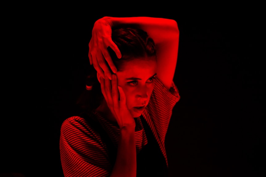 A brunette woman, wearing overalls, bathed in red light on stage, has one hand on top of her head, the other on her cheek