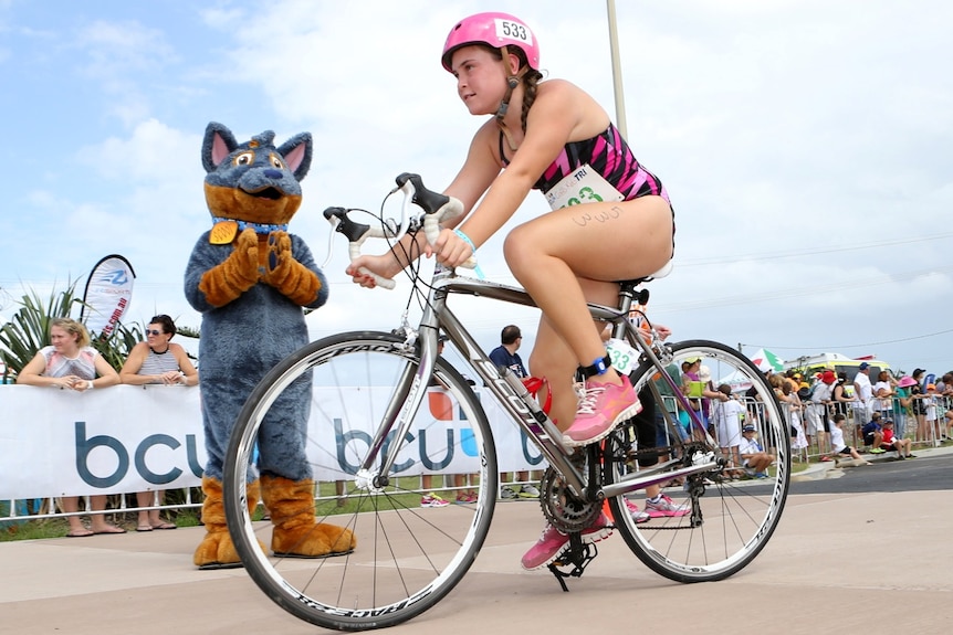 Girl in a pink helmet cycling past a group of onlookers while a mascot claps her on