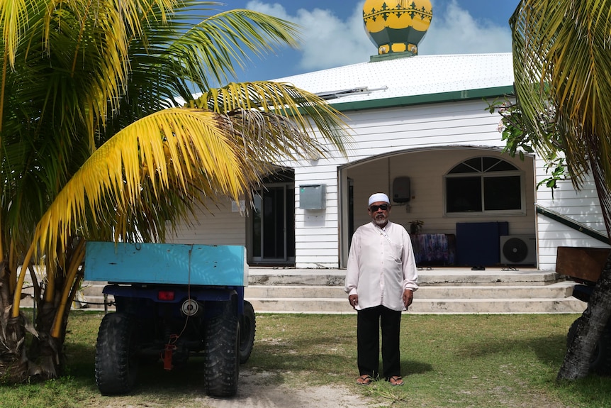 A man in formal white attire stands next to a 4wd buggy in front of a mosque with tropical palms.