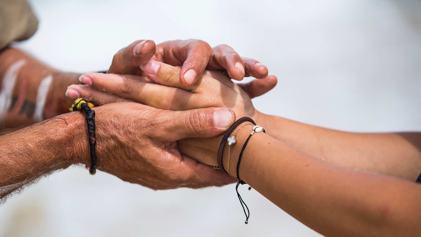 An older man’s hands with painted line markings grasp a pair of hands belonging to a younger person.