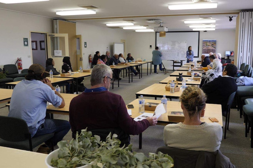 People sit at separate desks in a conference room with someone writing on a whiteboard at one end.