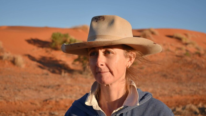 A woman in an old broadbrim hat stands in front of a bright orange sand dune in fading afternoon light.