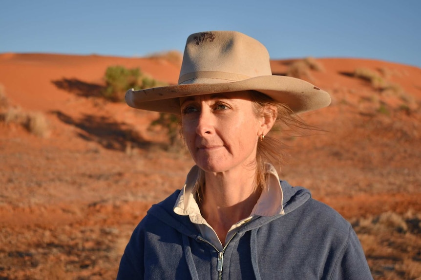 A woman in an old broadbrim hat stands in front of a bright orange sand dune in fading afternoon light.