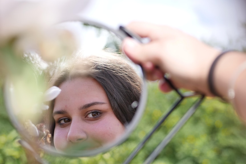 A teenage girl's reflection is seen in a small round mirror she holds up.