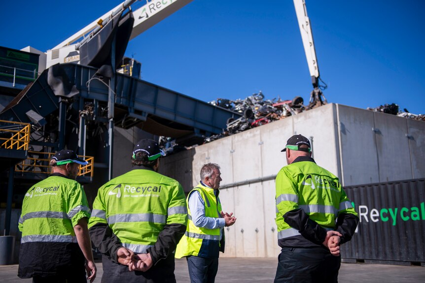 Four men in fluro vests face a scrap metal shredder and crane in the background.