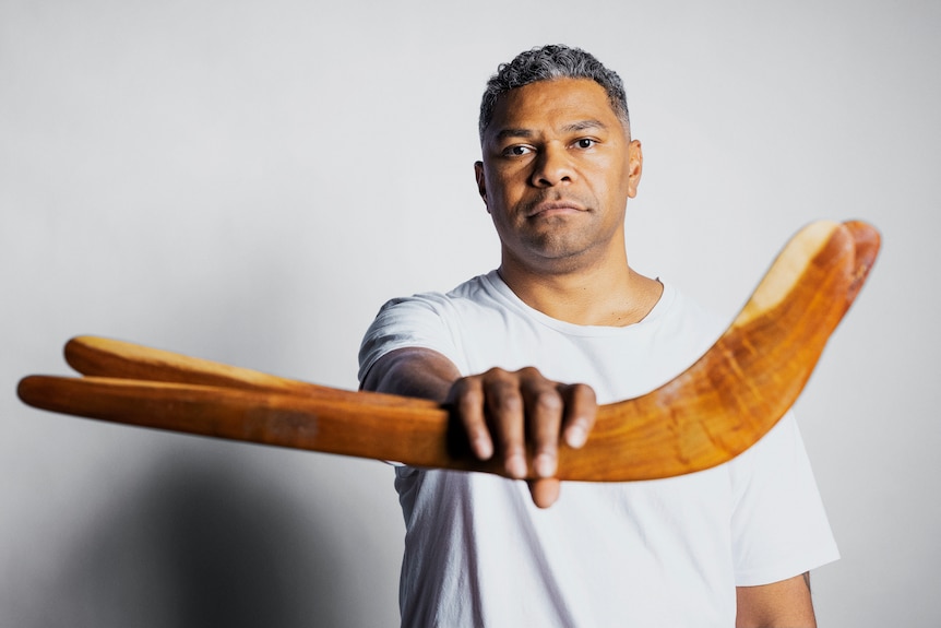 A dark-skinned man in a white t-shirt holds a boomerang in front of him.