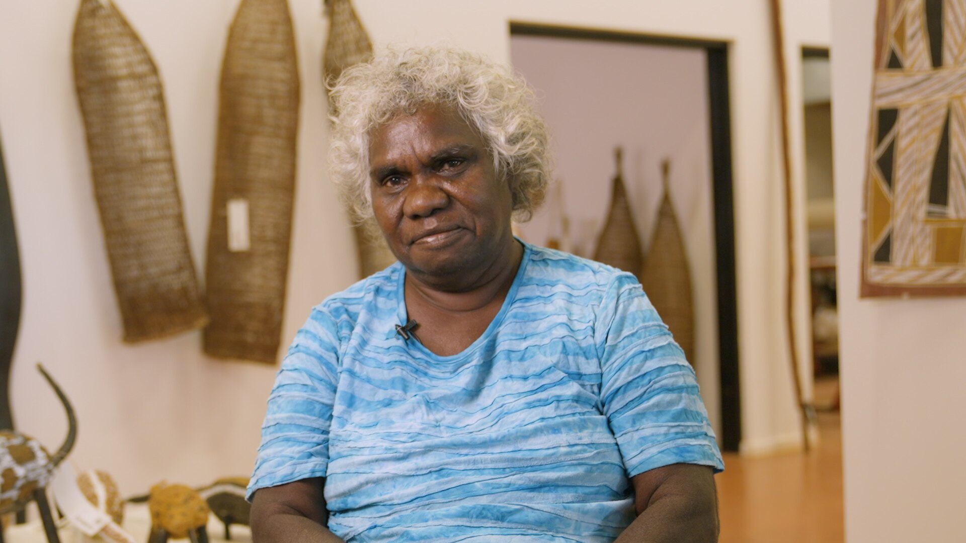 Doreen Jinggabbarra, a Burarra woman in her 60s with short curly grey hair, wearing a bright blue shirt in a gallery.