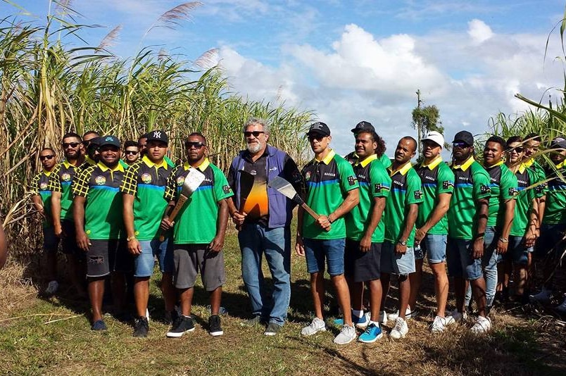 A group of young men dressed in green uniforms stand in a cane field holding cane cutters