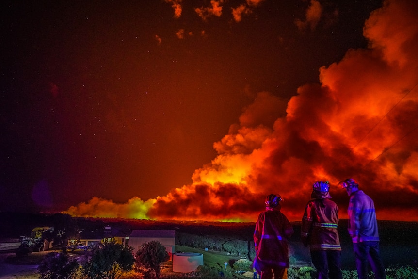 Three firefighters stand at night watching a blaze burn in the distance.