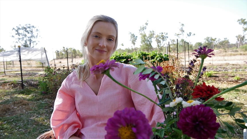 A young woman in a light coloured blouse is smiling, she has a colourful bouquet on the table in front & rows of flowers behind