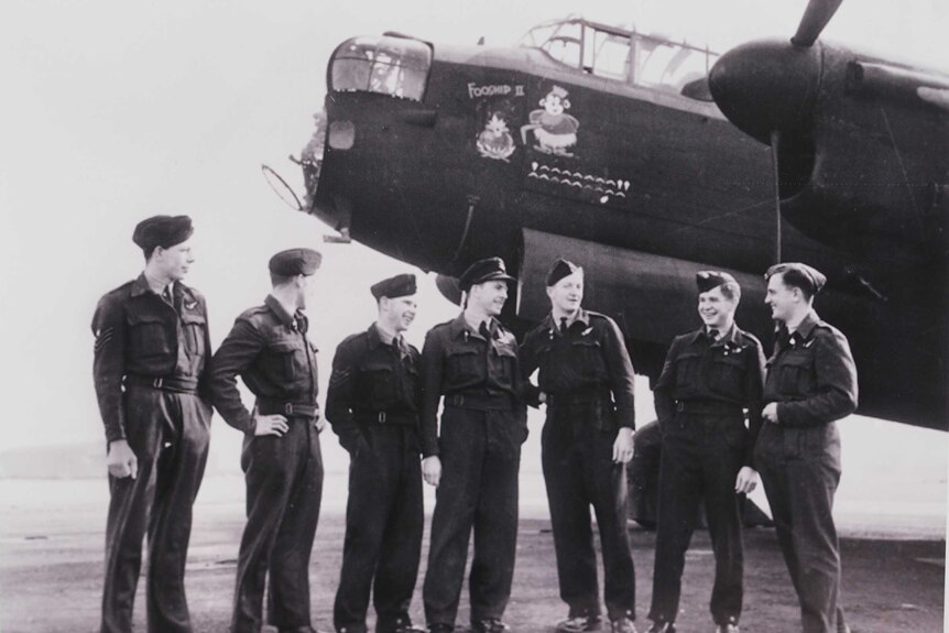 Black and white old photo of seven men in uniform standing in front of a plane.