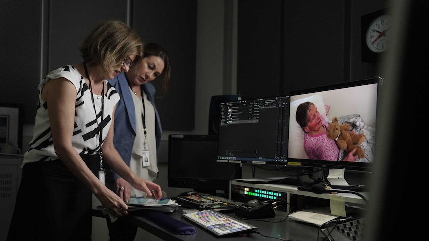 Puccini and Connolly standing in edit suite looking at script with image of elderly woman on screen.