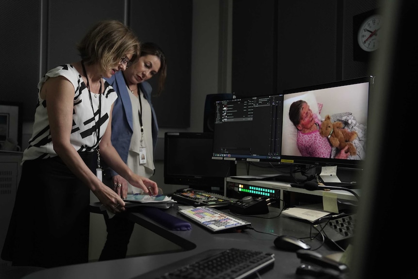 Puccini and Connolly standing in edit suite looking at script with image of elderly woman on screen.