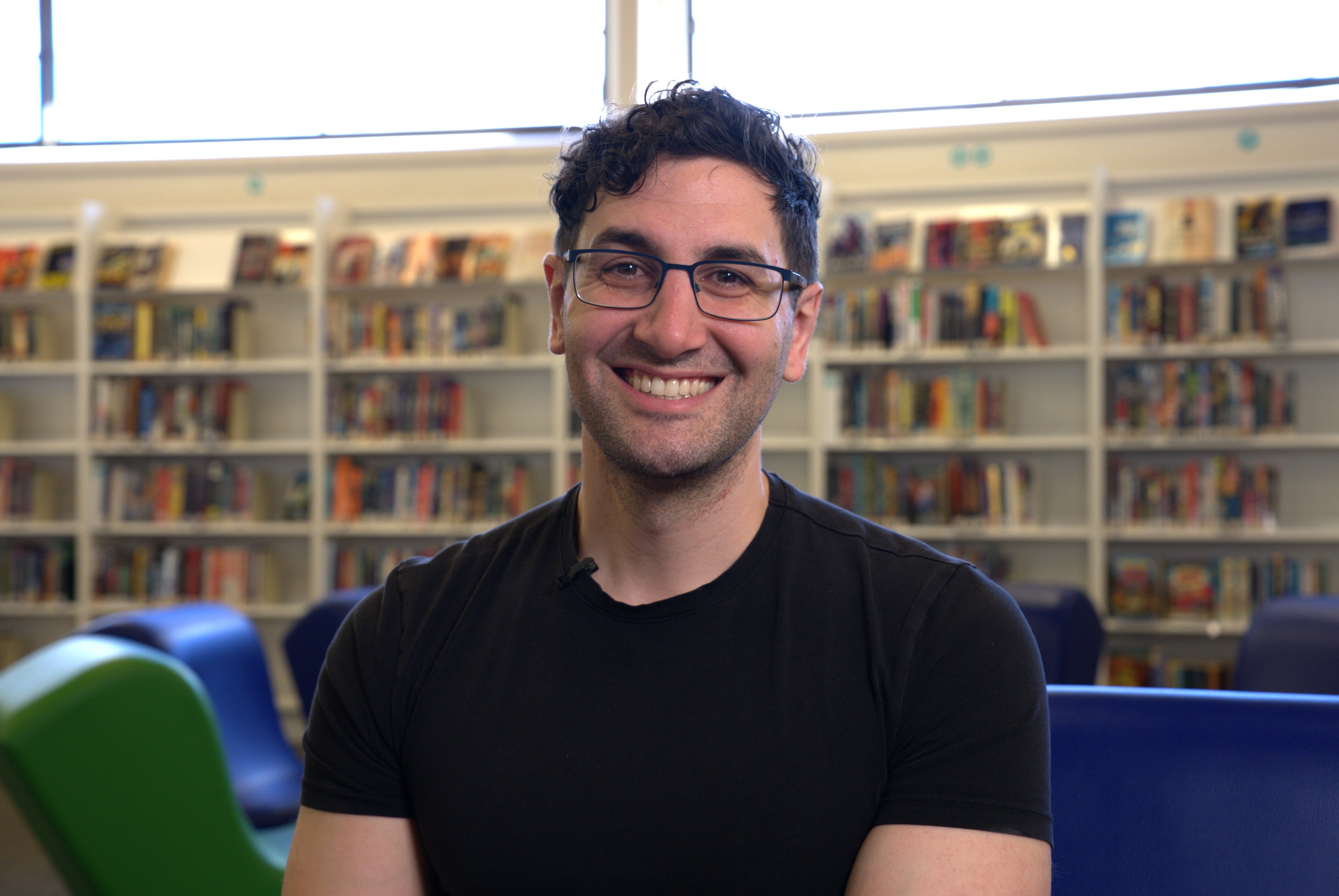 A man with dark hair, glasses and a black T-shirt smiles with bookshelves behind him.