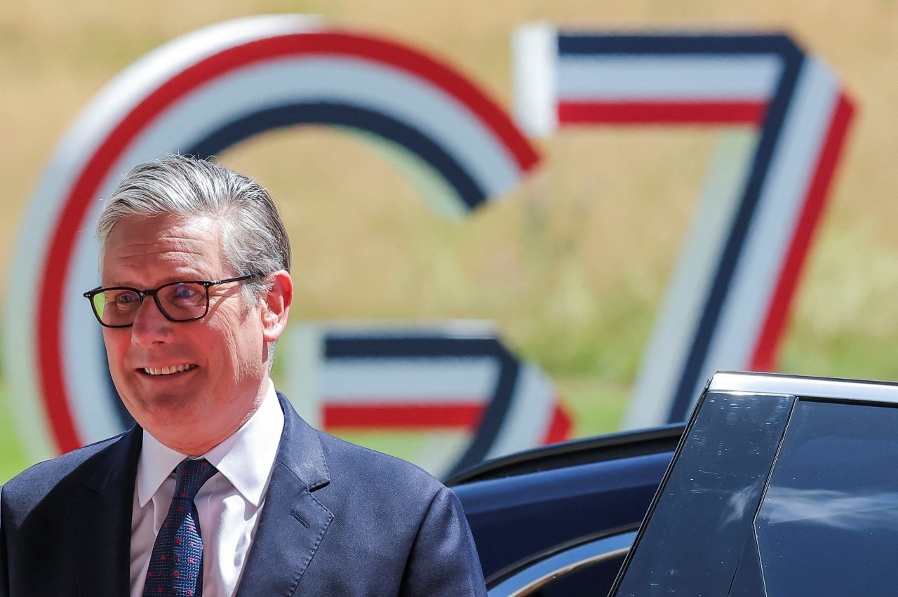 A man in a suit looking on and smiling. A large sign reading G7 can be seen in the background.