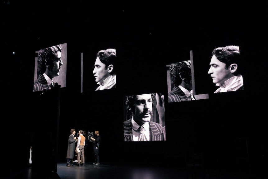 Two well-dressed men stand face-to-face on a stage. Close-ups of their faces are seen on five large hanging screens.