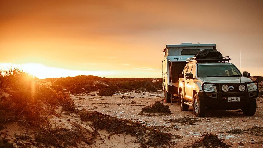 A caravan attached to a white four-wheel drive rests on brown sand with a sun on the horizon.