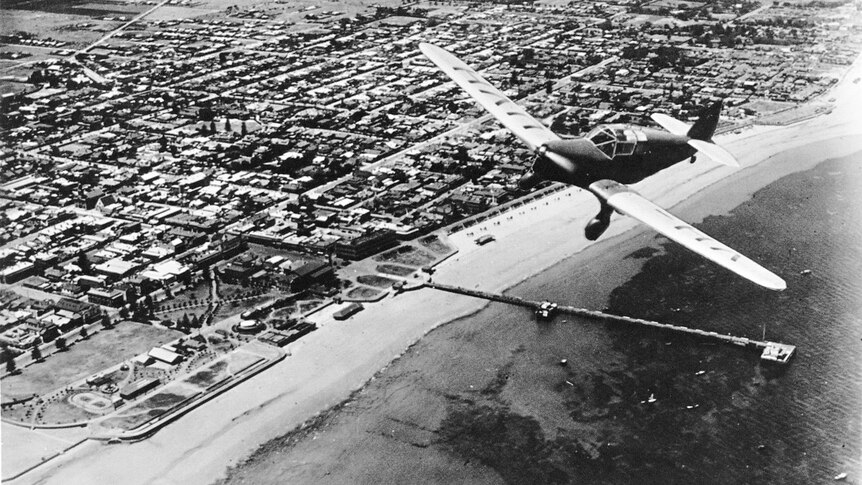 A black and white historical photo of an aeroplane flying over a town with ocean next to it