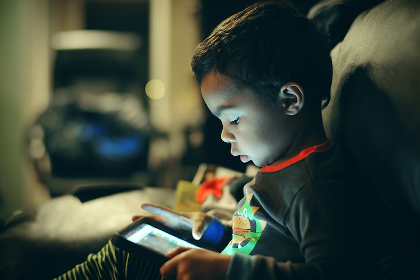 A child's face is illuminated by the iPad on their lap, as they tap the screen.