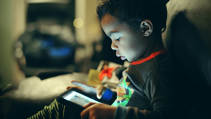 A child's face is illuminated by the iPad on their lap, as they tap the screen.